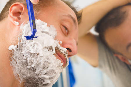 Man with lots of shaving foam on his face standing in bathroom, looking at mirror, shaving his face beard. Skincareの写真素材