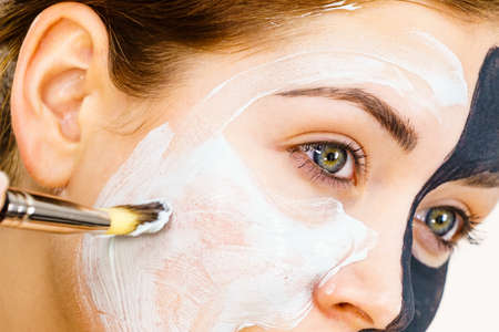 Woman with clay carbo black mask on half face applying white mud to clean skin. Girl taking care of...