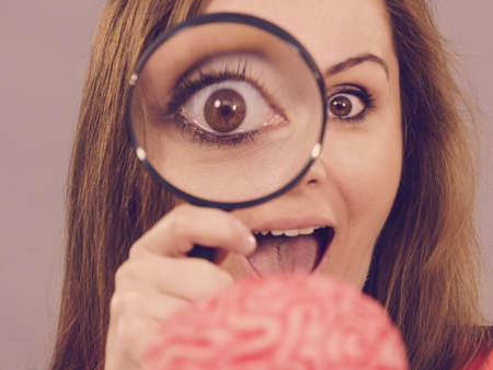 Close up of funny woman holding magnifying glass investigating something and looking closely at fake brain, examing intelligence.の写真素材