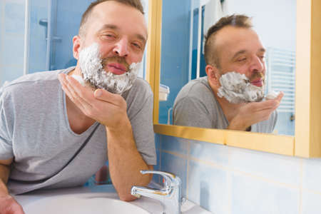 Man applying shaving foam cream on his face, standing in bathroom, looking at mirror, preparing to shave his face beard. Skincareの写真素材