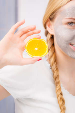 Facial dry skin and body care, complexion treatment at home concept. Happy young woman having grey mud mask on her face holding orange pieceの写真素材