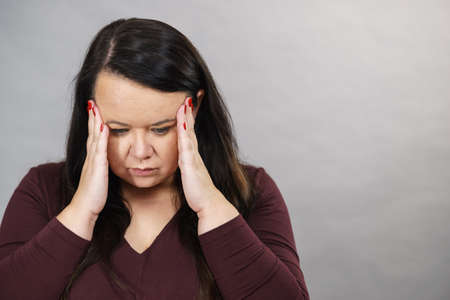 Headache, migraine and stress. Worried plus size adult upset woman suffering from head pain, studio shot on gray.の写真素材