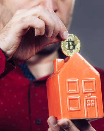Man paying his house rent using cryptocurrency. Guy holding model of red home and bitcoin coin.の写真素材