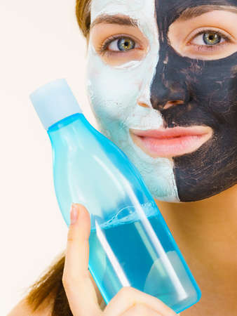 Woman with clay carbo black mask on one half face and white mud on second half. Girl removing cosmetic scrab with lotion. Female taking care of oily complexion. Skincare.の写真素材