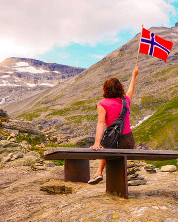 Tourist woman holds norwegian flag, enjoying nature landscape from Trollstigen viewpoint area. Trolls Path mountain road in Norway. Touristic attraction.の写真素材