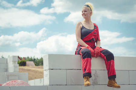 Woman wearing workwear on construction site. Female takes break from work, sitting on wall. Partially built new house early stage. Industry.の写真素材