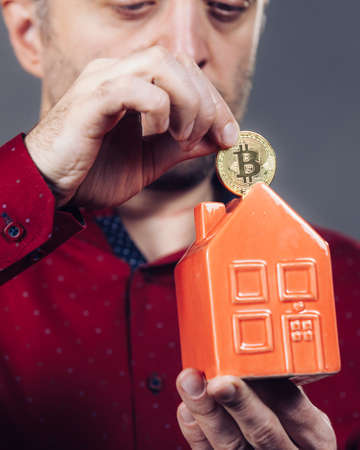 Man paying his house rent using cryptocurrency. Guy holding model of red home and bitcoin coin.