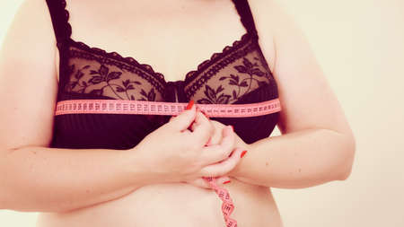 Plus size fat woman wearing black bra using tape measure to check the measurements of her big chest breasts. Closeup part of female body. Bosom, brafitting and underwear concept.の写真素材