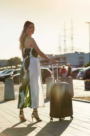 Fashionable woman arriving to new city wearing long dress and high heels, holding her suitcase on wheels admiring town after arrival.の写真素材