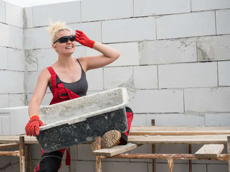 Woman wearing workwear working on construction site. Mixing cement in bowl preparing mortar. Partially built new home early stage. Industry.の写真素材