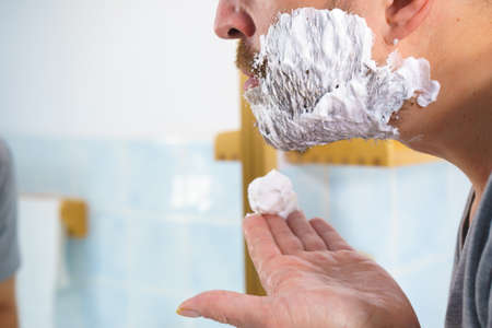 Man applying shaving foam cream on his face, standing in bathroom, looking at mirror, preparing to shave his face beard. Skincareの写真素材