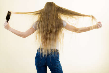 Blonde woman with brush combing her very long hair. Teenage girl with bad hair care. Back viewの写真素材