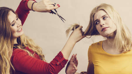 Hairstyle and haircut. Young female barber holding scissors tool ready to trimming hair her friends. Two girls creating new hairdo coiffureの写真素材