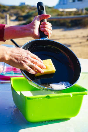 Mature woman washing up dishes in bowl on fresh air. Dishwashing outdoor on camping site, sea shoreの写真素材