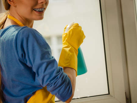 Young woman in yellow gloves cleaning window with blue rag and spray detergent. Spring cleanup, housework conceptの写真素材
