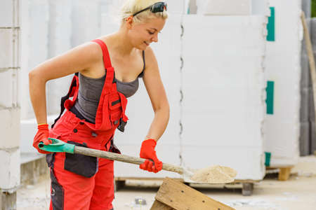 Woman working hard on construction site, using shovel digging sand soil. Partially built new home...