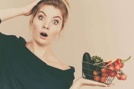 Adult woman do not like to eat raw food, questioning healthy lifestyle recommendations, origin vegetagles. Female holding small shopping basket with products, displeased shocked face expressionの写真素材