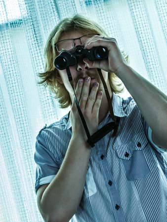 Young adult man spying at his neighbors, guy using binoculars looking through window.の写真素材