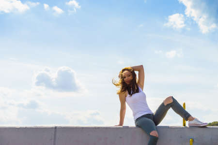 Trendy fashionable woman relaxing outdoor wearing casual footwear white sneakers and hole trousers. Female enjoy sunlight against blue sky.の写真素材