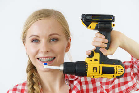 Funny teenage woman wearing braces holding drill about to brush her teeth with electric toothbrush...