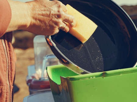 Mature woman washing up dishes in bowl on fresh air. Dishwashing outdoor on camping site, sea shoreの写真素材