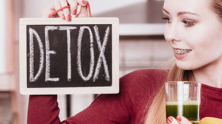 Drinks good for health, diet breakfast concept. Young woman in kitchen holding green healthy vegetable smoothie juice glass next to detox sign board.の写真素材