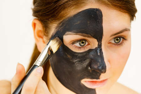 Skin care. Female applying black purifying mud mask, cosmetic healing clay to half face, using brush. Beauty and wellness. Spa and acne treatment.の写真素材
