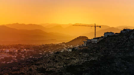 Sunset over spanish coastal landscape with construction site with crane industrial equipment on hill. Building activity.の写真素材
