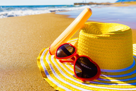 Sun protection accessories on sea shore. Red sunglasses, yellow straw hat and sunscreen cream bottle on sandy beach. Summer and holidays.の写真素材