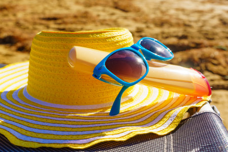 Sun protection accessories on sea shore. Blue sunglasses, yellow straw hat and sunscreen lotion on sandy beach. Summer and holidays.の写真素材