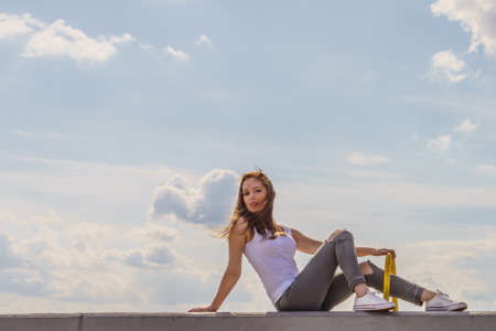 Young pretty fashion model woman sitting on concrete wall wearing white tank top and olive green...