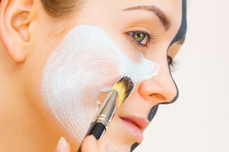 Woman with clay carbo black mask on half face applying white mud to clean skin. Girl taking care of oily complexion. Beauty procedures. Skincare.の写真素材