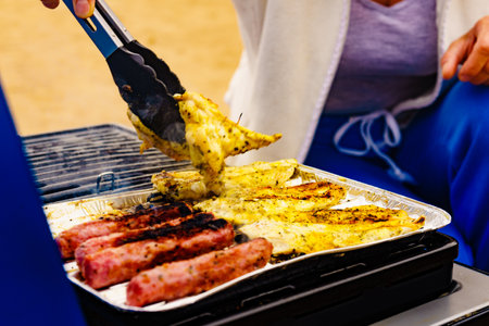 Hand of woman grilling chicken meat and sausages on barbecue gas grill. Barbeque dinner outdoor.の写真素材
