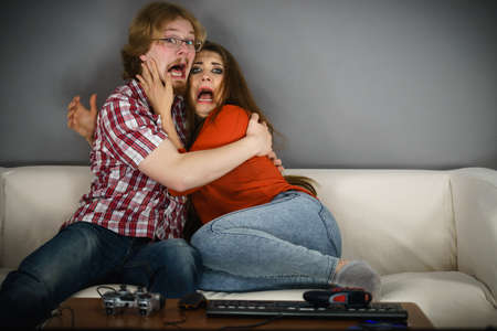 Funny couple sitting on sofa watching horror movie being scared hugging each other.の写真素材