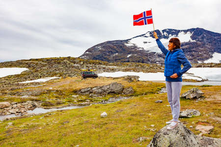 Woman holds norwegian flag in mountains, van with canoe on top roof in distance. National tourist scenic route 55 Sognefjellet, Norwayの写真素材