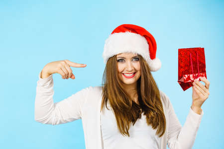 Smiling woman is happy to give Christmas gifts. Female wearing santa claus hat points to present red...