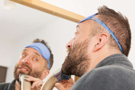 Bearded man looking at himself in mirror trimmng, shaving his beard using electric timmer razor. Guy taking care of facial hair, wearing sleepy eyemask.の写真素材