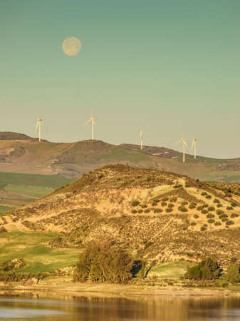 Wind turbines on hills. Full moon on sky at morning. Power generator farm for clean renewable electric energy production near Ardales, province Malaga, Andalusia Spain.の写真素材