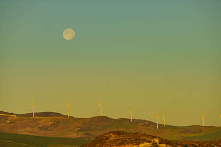 Wind turbines on hills. Full moon on sky at morning. Power generator farm for clean renewable electric energy production near Ardales, province Malaga, Andalusia Spain.の写真素材