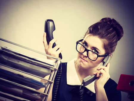 Mobbing at work, bad job relations concept. Angry mad bossy businesswoman talking on phone sitting working at desk full off documents in binders.の写真素材