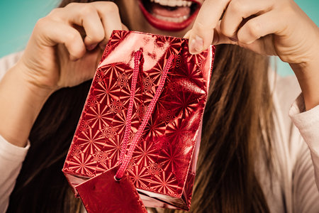 Happy woman wearing santa claus hat looks into christmas red gift bag, on blueの写真素材