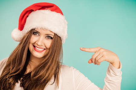 Young brunette smiling woman in christmas santa hat pointing with finger to her face, on blueの写真素材