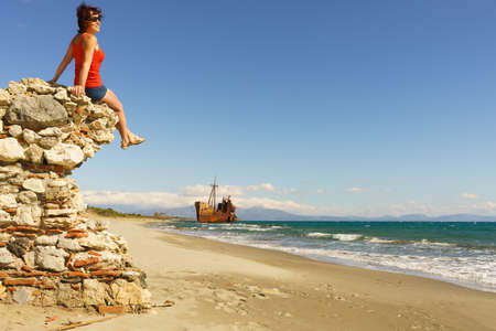 Travel freedom. Mature tourist woman on beach enjoying summer vacation. An old abandoned shipwreck, wrecked boat in the backgroundの写真素材