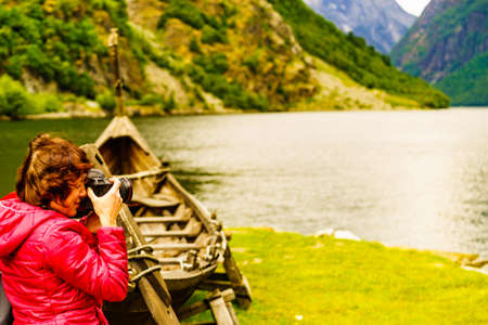 Female tourist near old wooden viking boat on norwegian nature fjord shore, taking photo with camera. Tourism and traveling conceptの写真素材