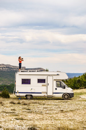 Woman enjoying trip with motor home, standing on roof of camper vehicle, holds camera, taking travel picture from mountain nature. Happiness, vacation and traveling.の写真素材