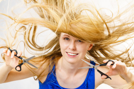 Girl with blowing long blonde hair holding scissors, showing work tools, normal and chunking shears. Making hairstyle, new hairdo. Haircare concept.の写真素材