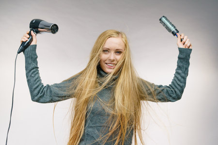 Female with blowing messy hair holding accessories hairdryer and brush. Blonde woman styling her very long disobedient hair.の写真素材