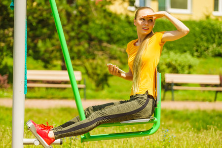 Young woman working out in the outdoor gym. Girl holding mobile phone while doing exercises on street machine.の写真素材