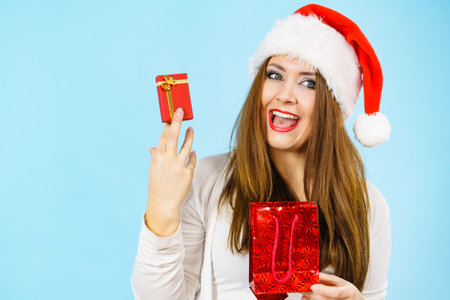 Christmas Gifts. Happy woman wearing santa claus hat holding present red bag and small gift box with jewelery, on blueの写真素材