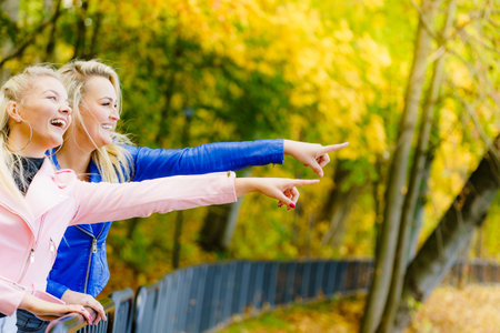 Two blonde women best friends wearing fashionable outfit, pink and blue ramones jackets, walking in autumn park.の写真素材